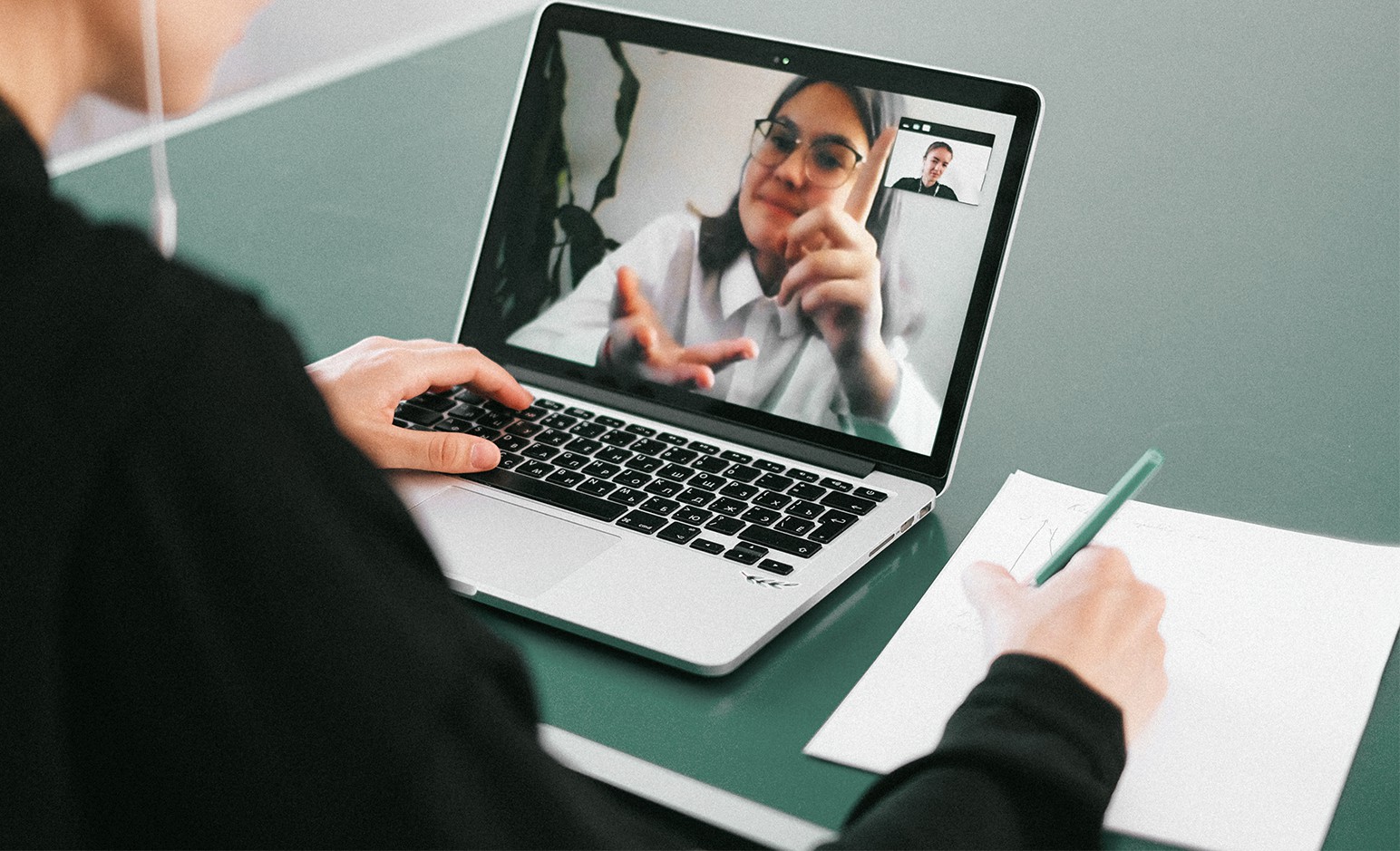 - Schulterblick in eine Remote-Interview-Session: Eine junge Frau ist auf dem Bildschirm eines Laptops zu sehen, während die Interviewende Person auf einem Notizblock mitschreibt.