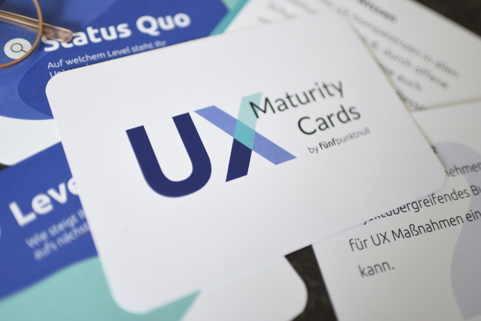 - Mehrere Karten des UX-Maturity Kartensets liegen verteilt. Mittig befindet sich eine Karte mit der Aufschrift: UX Maturity Cards by fünfpunktnull.