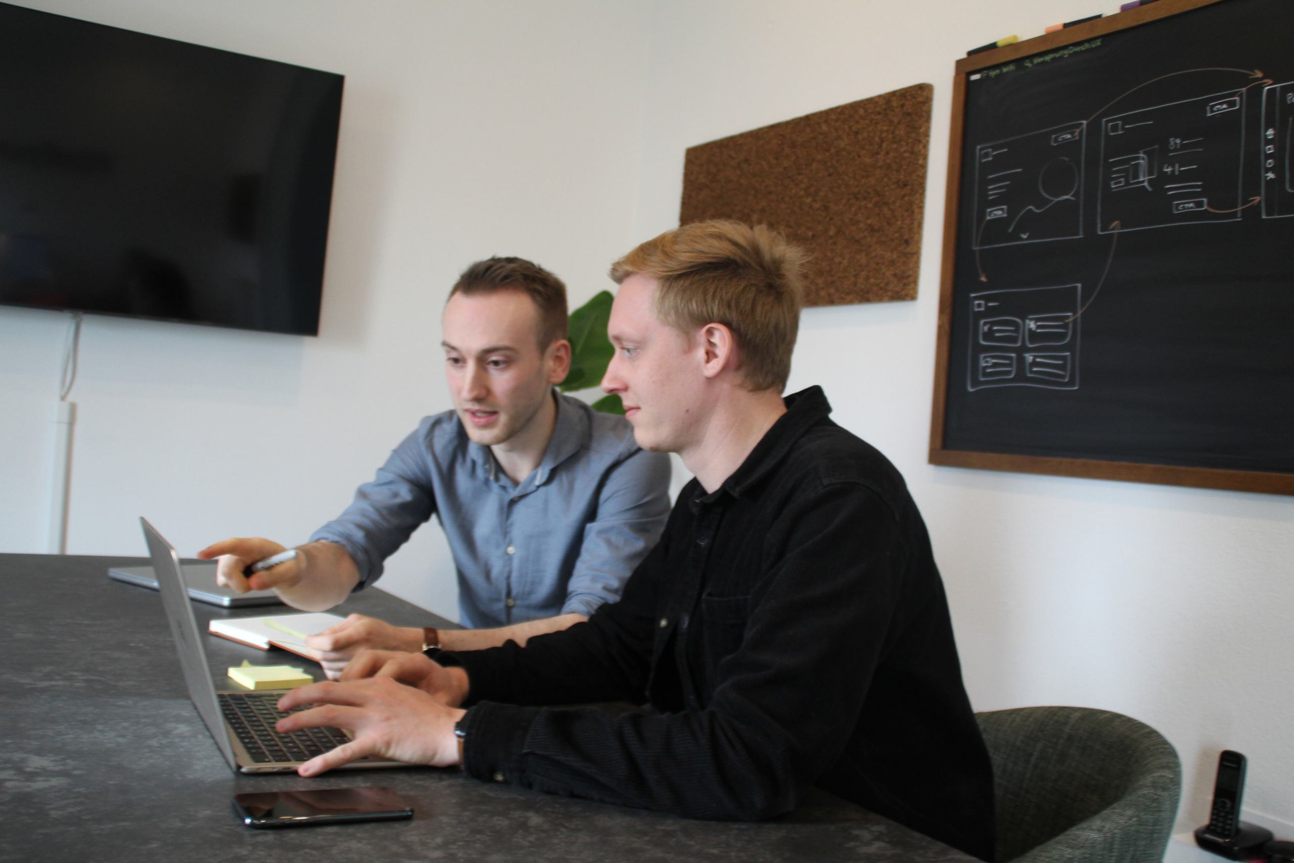 - Zwei Personen stehen zu einer schwarzen Tafel gewandt, auf der Notizen und Scribbles von User Interfaces zu sehen sind. Der Mann schreibt auf das Bord und schaut zur Frau, welche ein Post-it auf die Tafel klebt. 